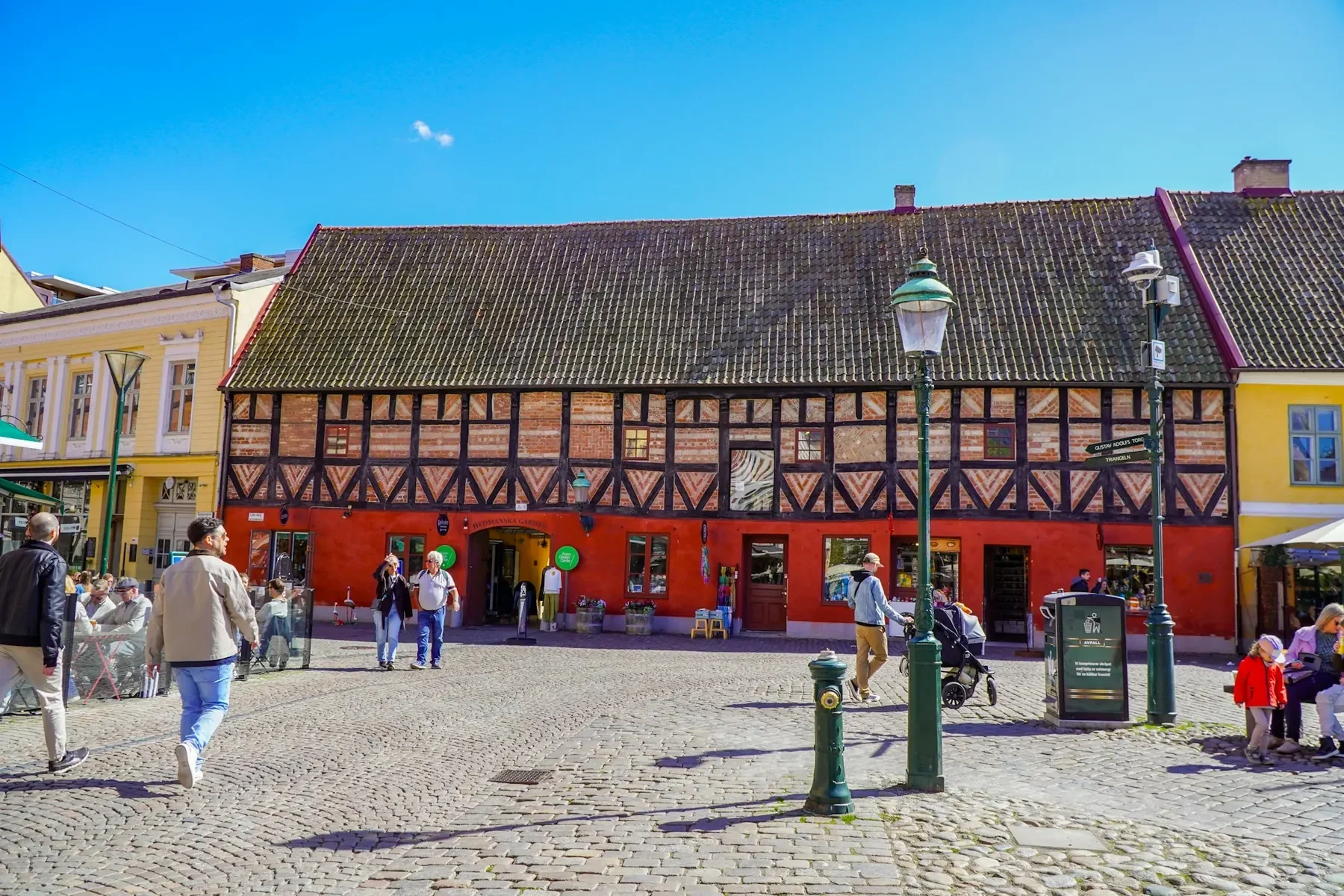 Lilla torg i Malmö en solig dag, med ett rött korsvirkeshus, gatlyktor och människor på kullerstenen.