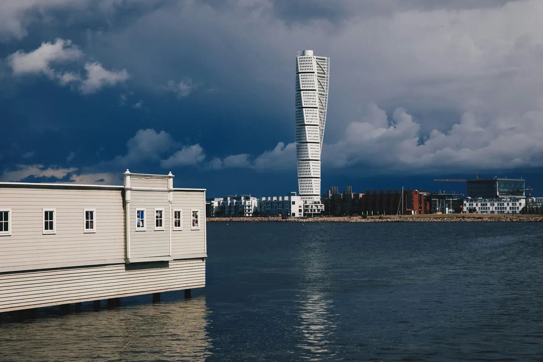 Turning Torso i Västra hamnen i Malmö under mörka moln, sett över vattnet med ett vitt kallbadhus i förgrunden.