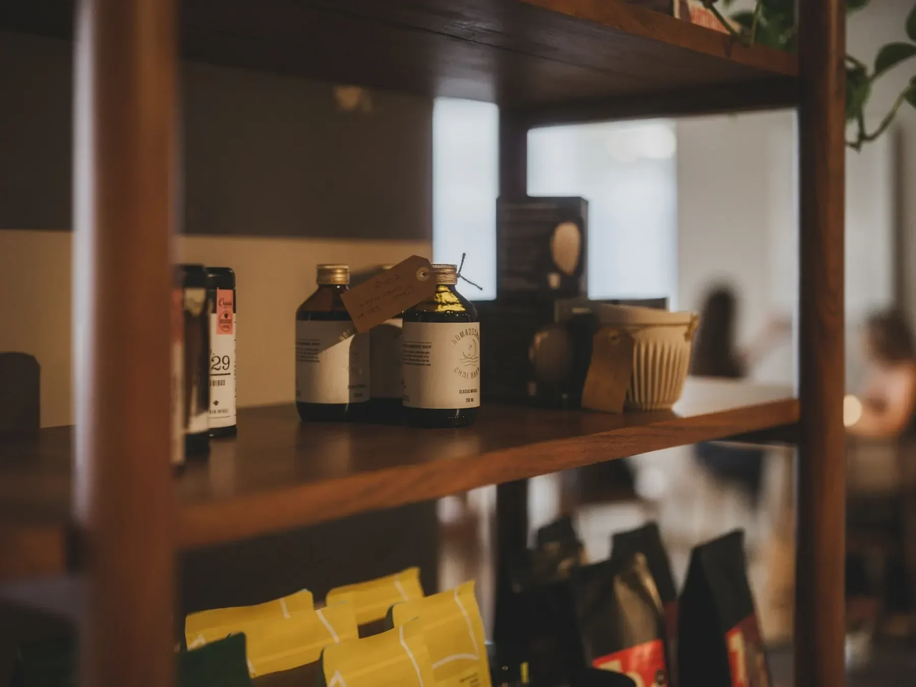 Trähylla i en liten butik med flaskor, kaffepaket och prislappar, mjukt suddig bakgrund.